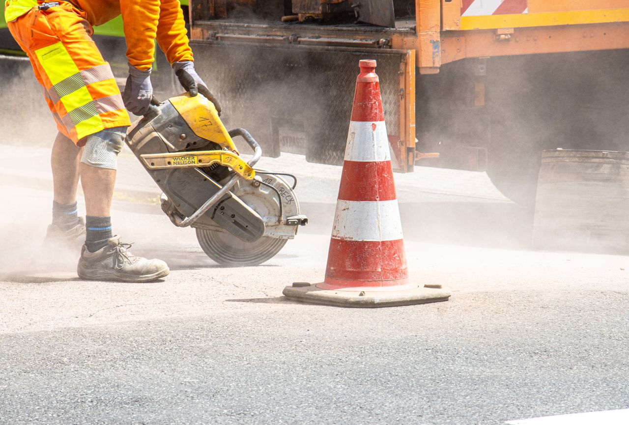 A construction worker cutting asphalt with a large saw next to a traffic cone on a street.