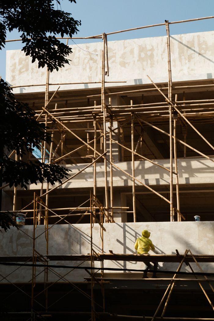 A worker in a yellow jacket climbs bamboo scaffolding at a construction site under bright daylight.